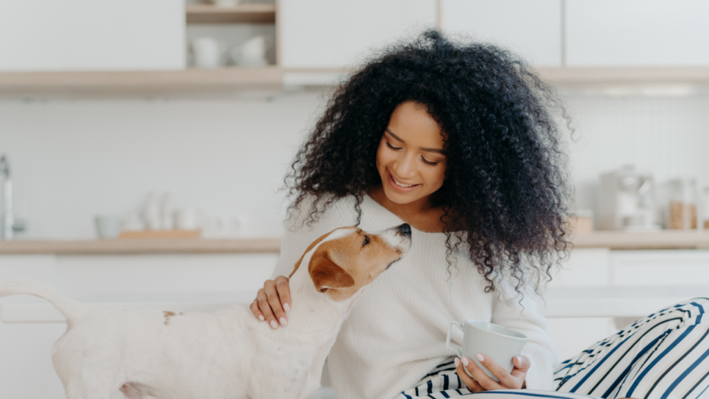 Woman with curly hair petting a small dog while holding a mug in a kitchen.
